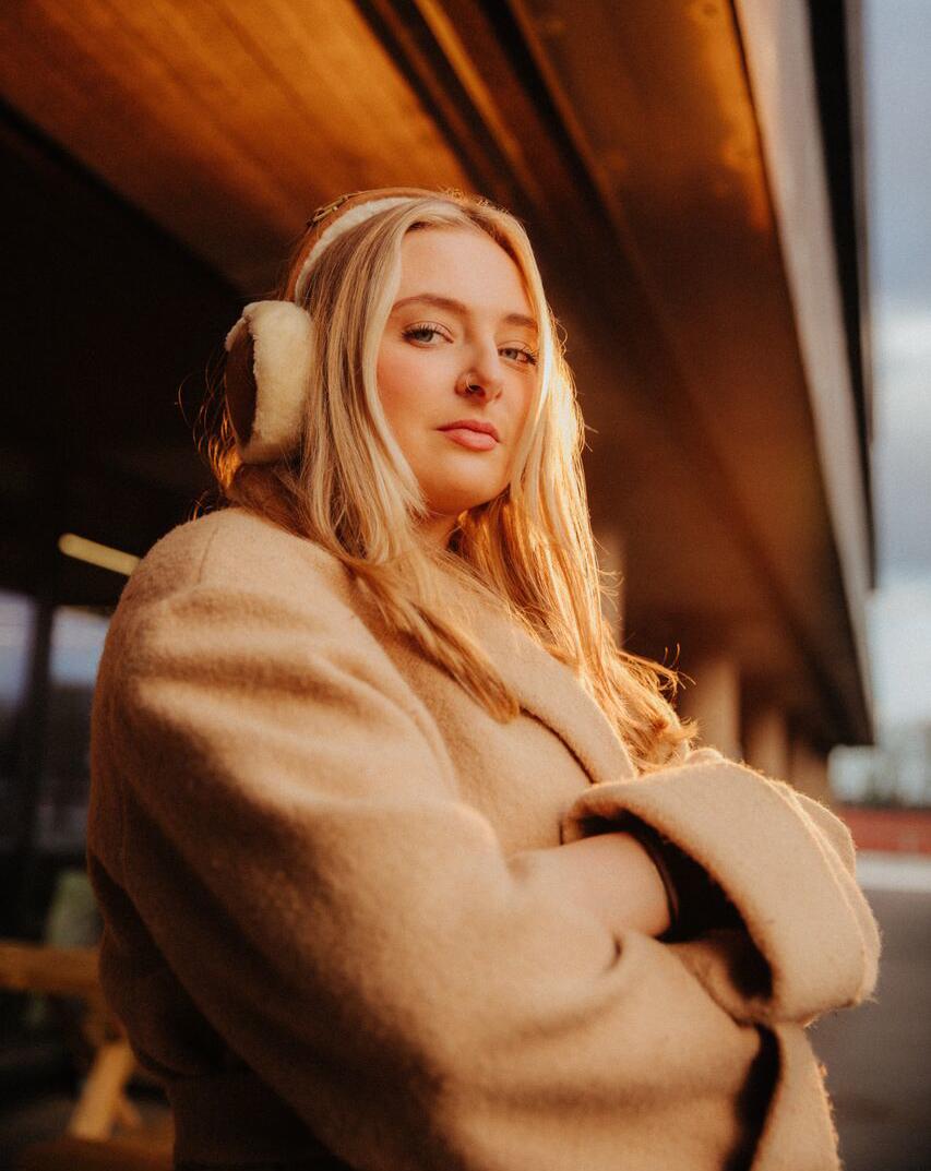 A female student in a beige coat with matching earmuffs stands confidently in dramatic sunlight outside the students' union