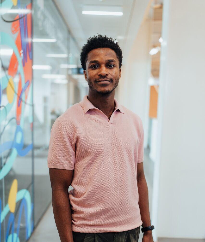 A male student in pink polo shirt stands confidently by patterned glass panels above the sports arena