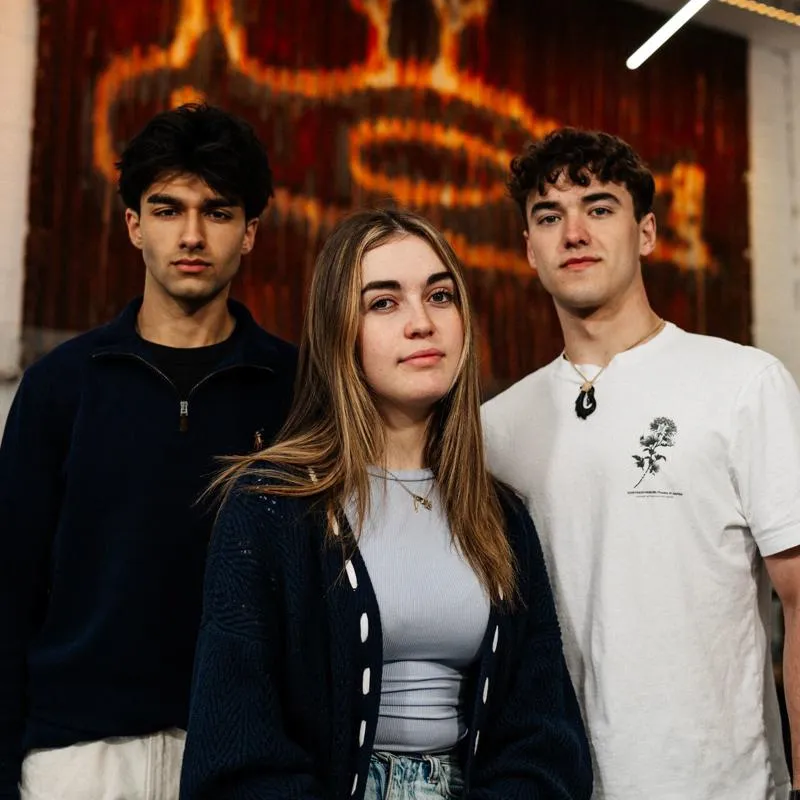 Three of Hull’s students stand confidently together in front of a rusted panel with Hull's iconic 'Dead Bod' artwork