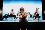 A music student tunes a violin while other musicians set up on a stage behind her