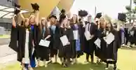 A group of new graduates stand on the lawn outside Connexin Live waving their hats and holding degree certificates.
