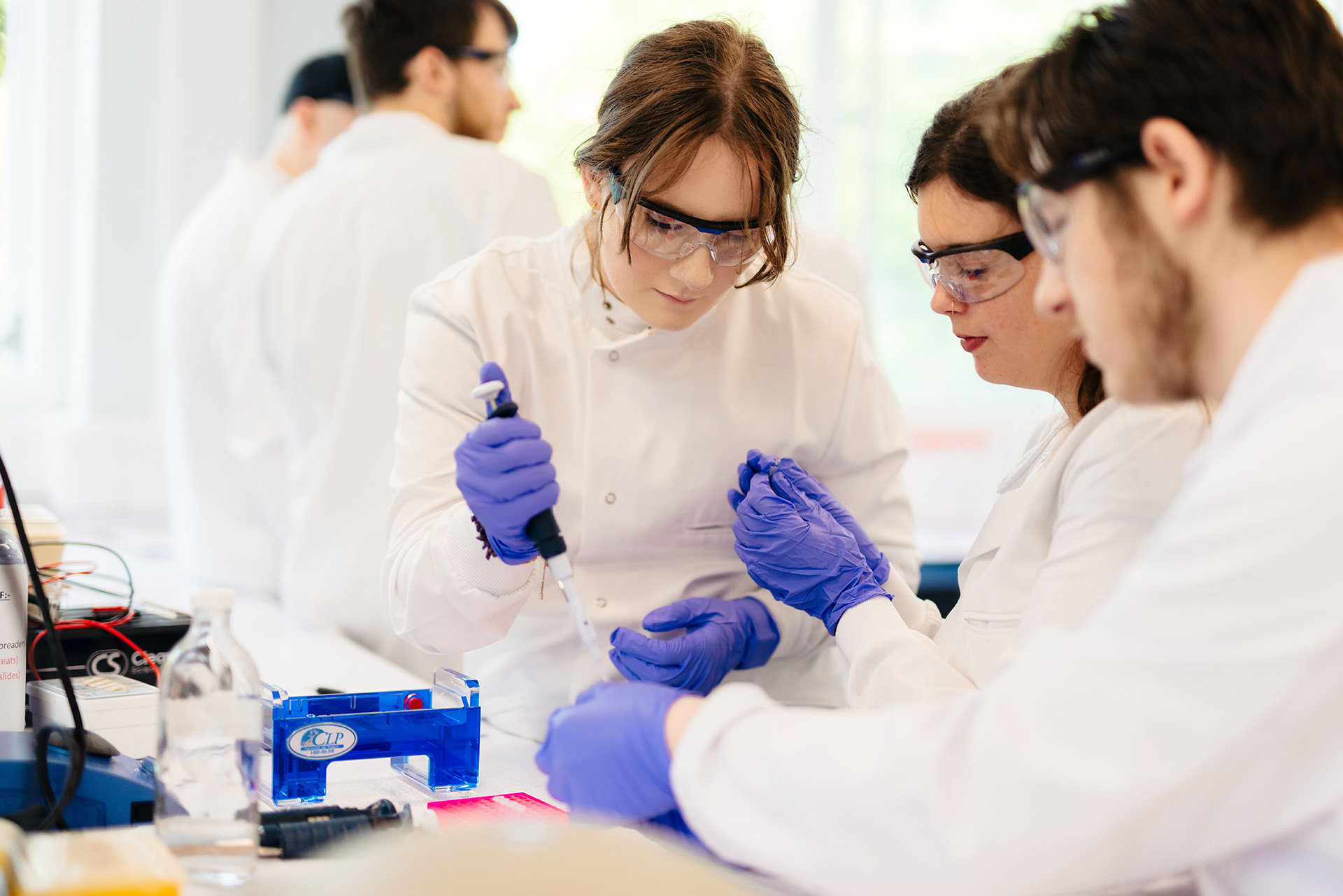 Students wearing white lab coats and PPE in a laboratory using a pipette