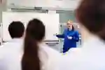 An instructor in a blue lab coat demonstrates on a whiteboard