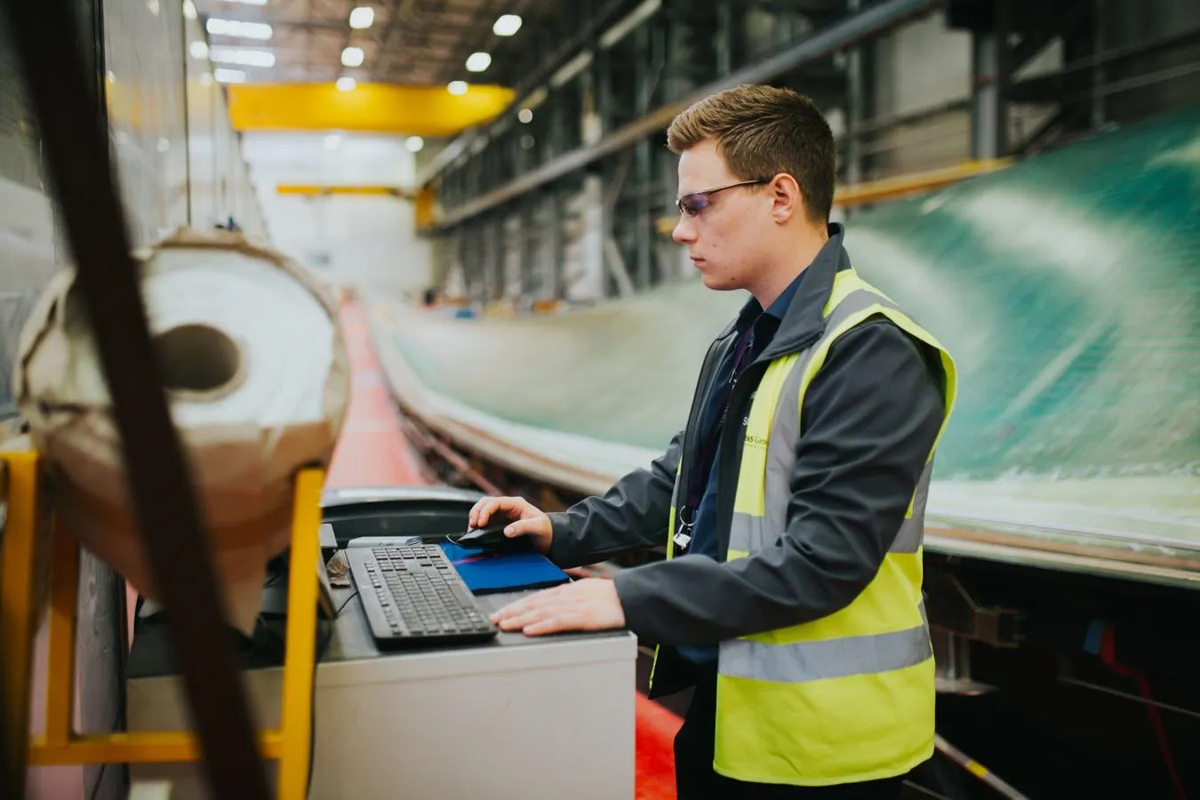 A student in hi-vis and protective goggles operates a computer in front of a large wind turbine blade