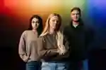 Three students stand confidently in front of prismatic light projected on to a wall behind them