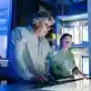 A lecturer gestures towards an item on a screen embedded in a table, surrounded by students in an engineering lab