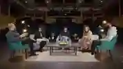 A group of people sit in comfortable chairs around a coffee table on a stage
